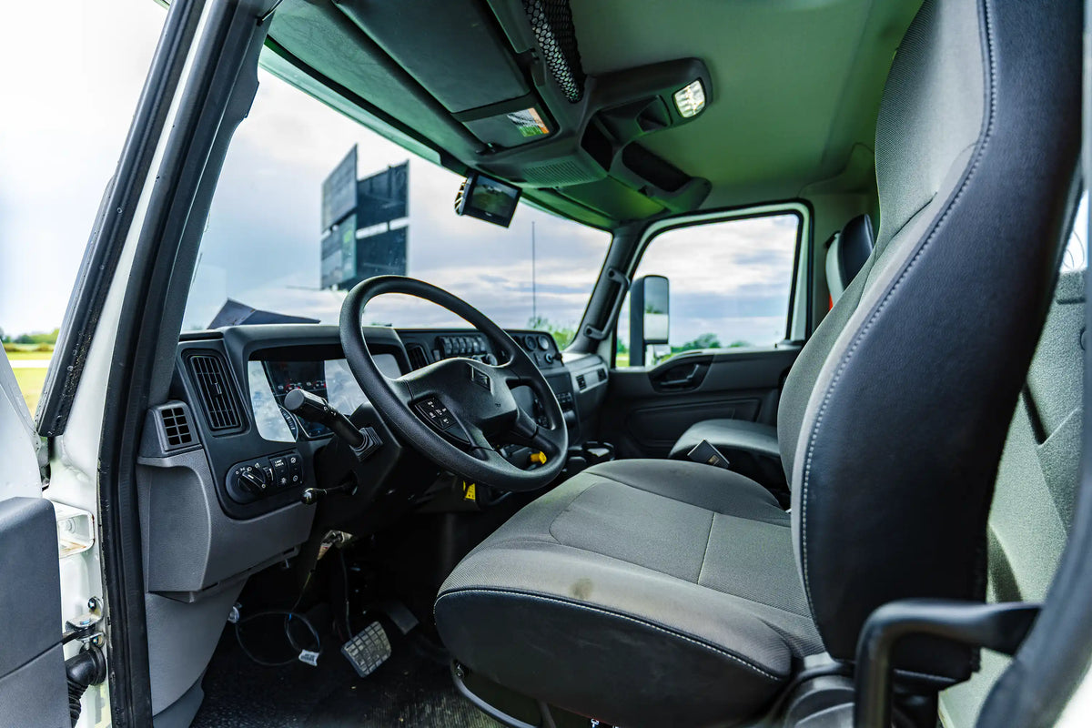 The driver-side interior truck cab of the 2026 International MV-607 1900 Gallon Vacuum Truck for sale from Arkson Truck Co, showing the driver's seat, steering wheel, dashboard, and side mirror against a bright clouded outdoor sky.