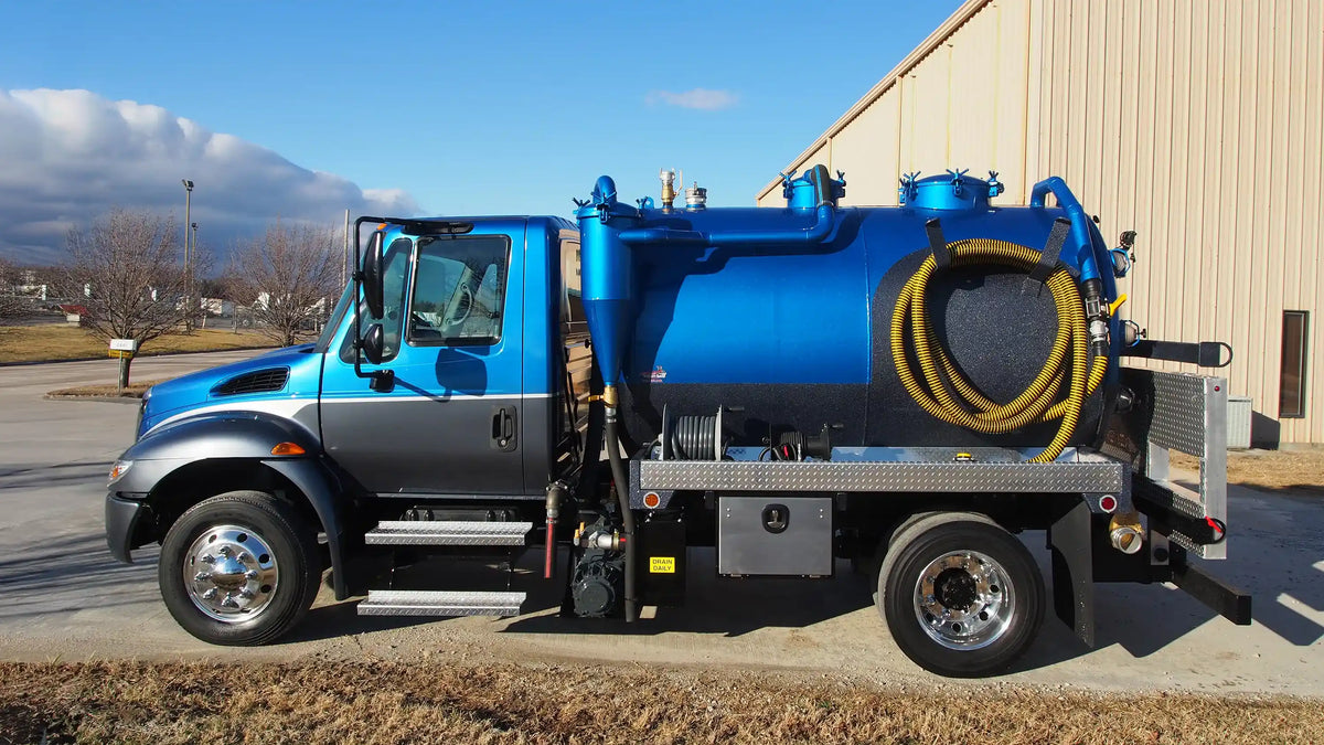 A blue and gray custom portable restroom truck with color matched vacuum tank for porta potty service applications parked outdoors beside a beige building under a blue sky.