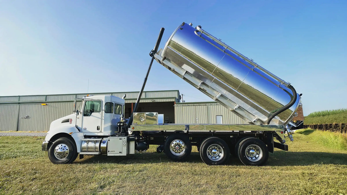 Industrial vacuum truck built by Arkson Truck Co, parked on the grass in front of a metal shop building with the debris body raised, under a blue sky.